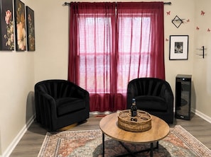 Un salon avec deux fauteuils noirs, une table basse ronde agrémentée d’un élément décoratif, un rideau rouge et une grande fenêtre.