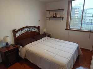 A bedroom with a bed, nightstands, a lamp, a window with blinds, and a shelf with decorative items. - Apartment in Lima with transfer included (Santiago de Surco)