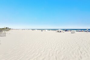 A wide sandy beach with a clear blue sky, a few beach umbrellas, and a distant view of the ocean. - Phoenix 10 Unit 1417 |  Next to Flora-Bama! | Beachfront | Pools | My Beach Getaways (Orange Beach)