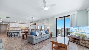 Amplia sala de estar con sofá, mesa de centro y mueble para televisor, una cocina con armarios blancos y un ventanal con vistas al océano.