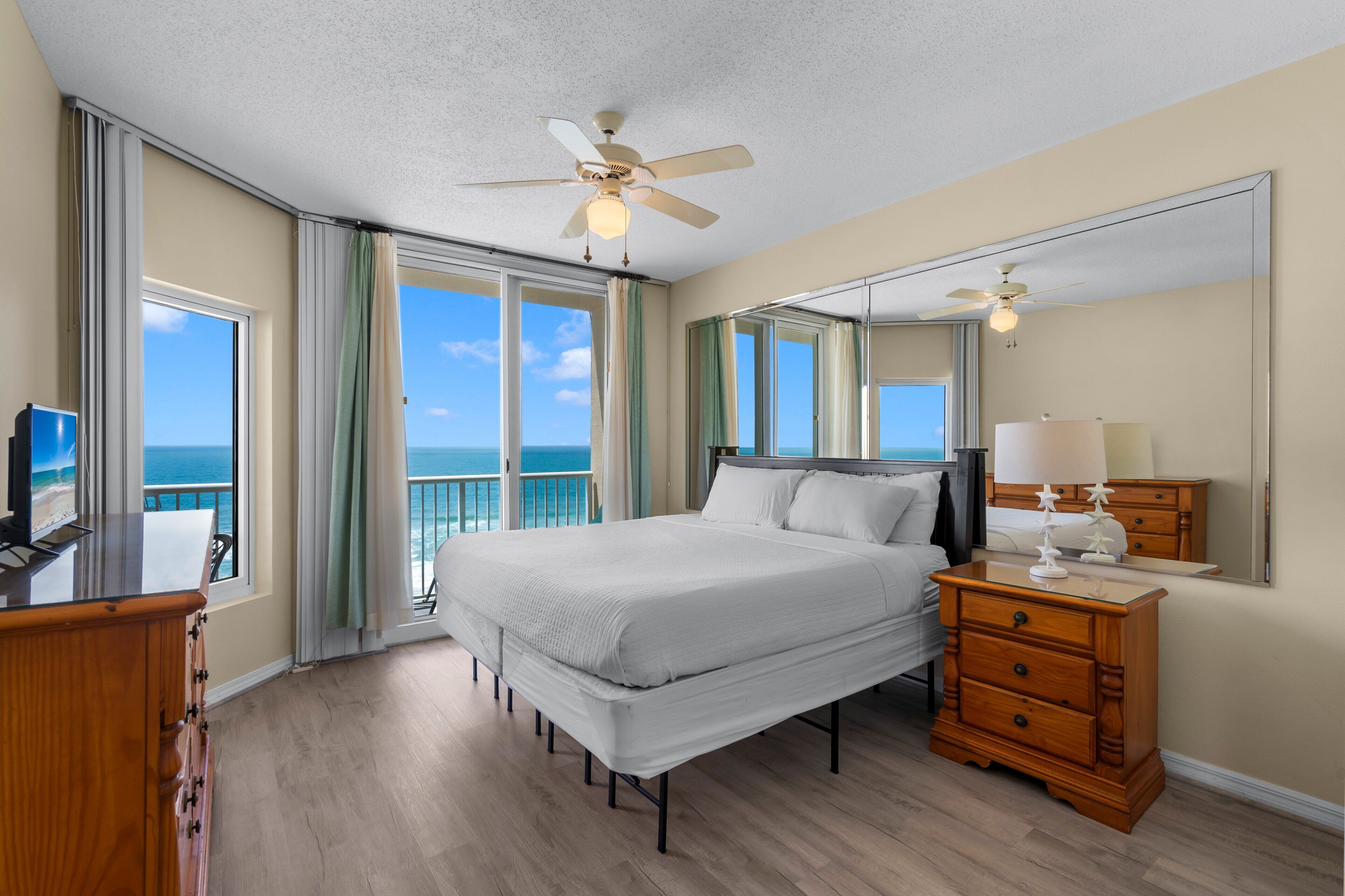 A hotel room with a bed, a dresser, a television, and a large window offering a view of the ocean.