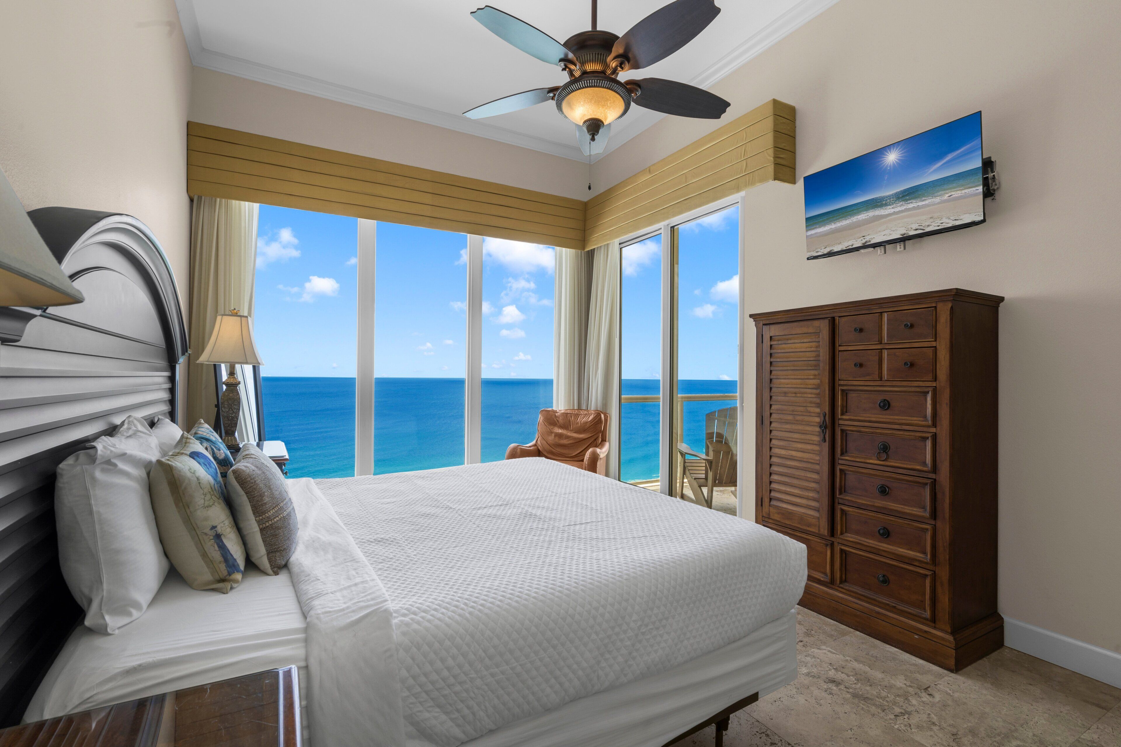 A hotel room with a bed, a wooden cabinet, a ceiling fan, and a large window offering a view of the ocean.