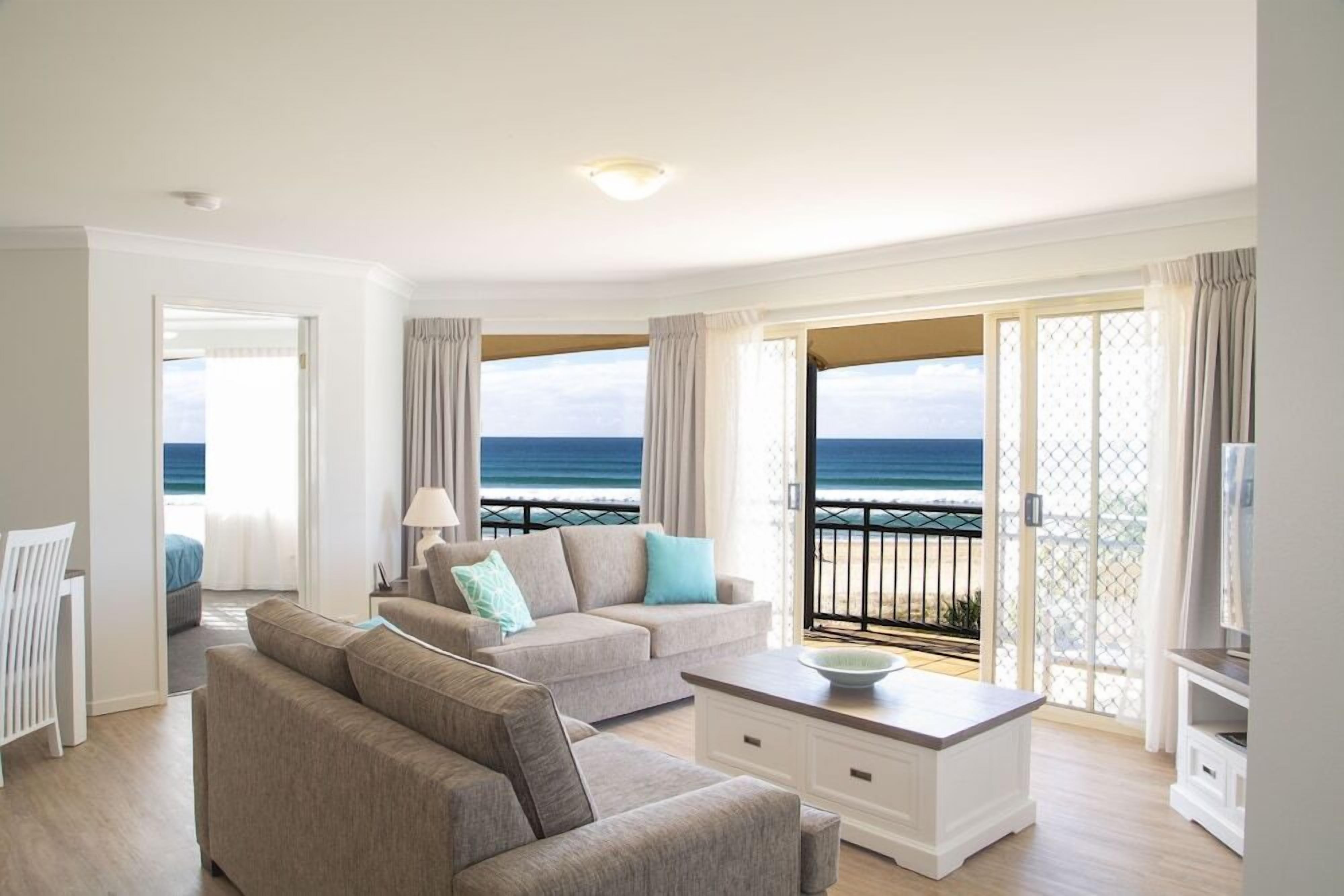 A modern living room with a sofa, a coffee table, and a TV stand. There is a balcony with a view of the sea.