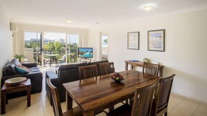A spacious living area with a dining table, sofas, and a balcony with a TV. - Golden Riviera (Gold Coast)