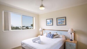 A hotel room with a large bed, two bedside tables, a ceiling fan, and a window with a view. - Golden Riviera (Gold Coast)