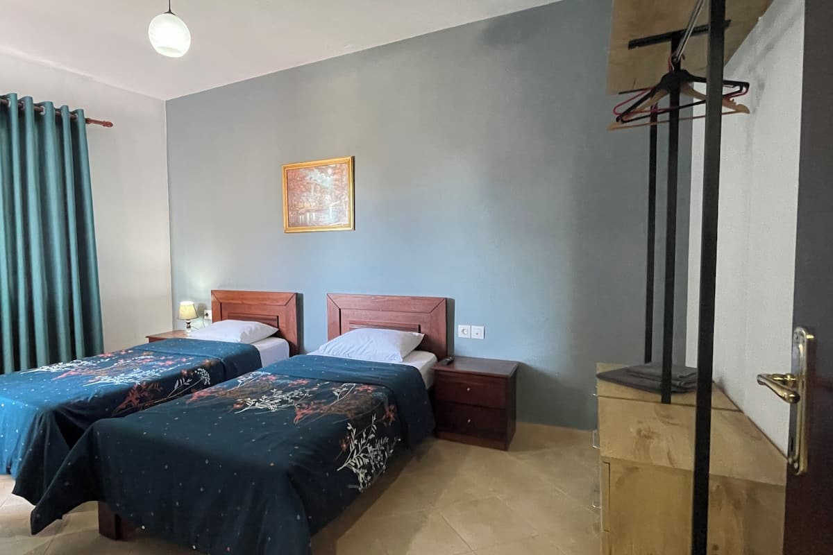 Una habitación con dos camas, un armario de madera, un perchero y un cuadro en la pared, además de una ventana con cortinas.