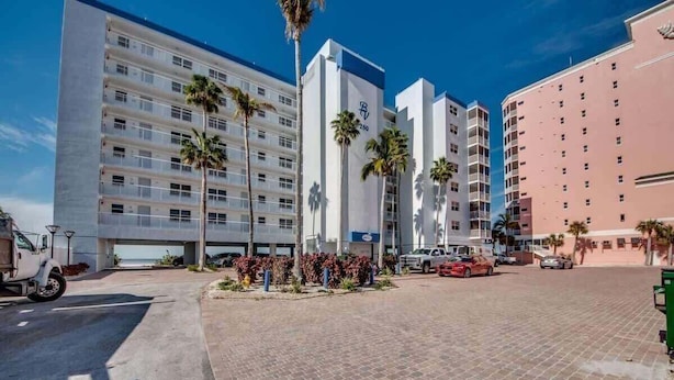 Een hotel met meerdere verdiepingen, een centraal binnenterrein, palmbomen en een parkeerplaats.