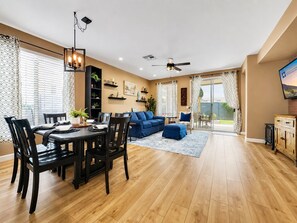 Ein modernes Wohnzimmer mit Essbereich, einem blauen Sofa, einem Deckenventilator und einem an der Wand befestigten Fernseher.