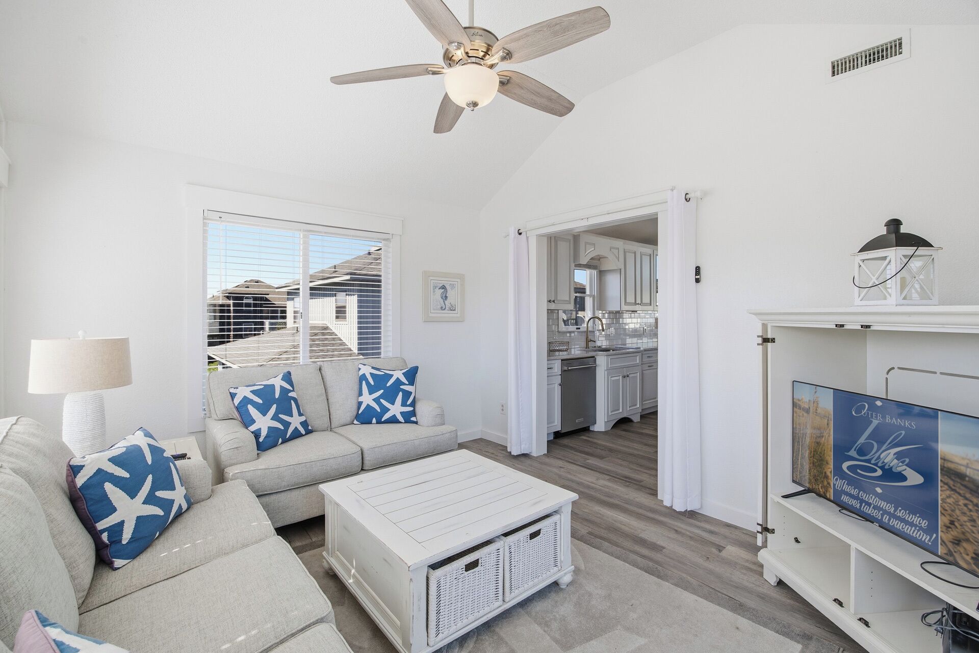 A living room with a ceiling fan, a sofa with star-patterned pillows, a white coffee table, and a kitchen in the background.