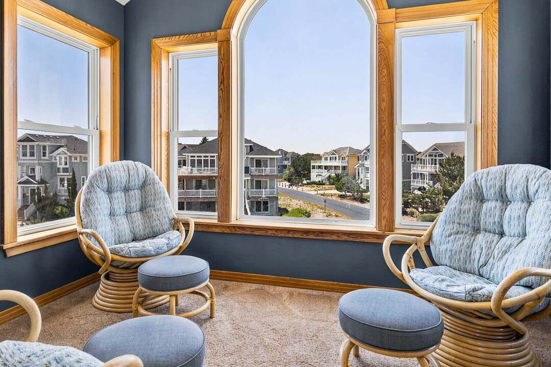 A room with three wicker chairs and ottomans, large windows with wooden frames, and a view of residential buildings.