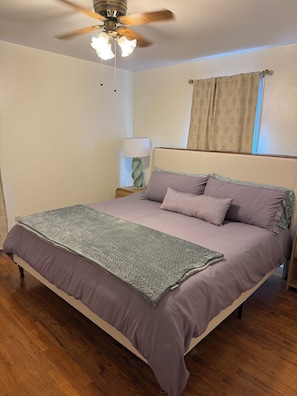 A bedroom with a bed, a ceiling fan, a nightstand with a lamp, and a window with curtains.