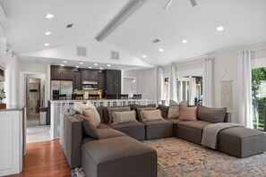 A spacious living area with a large sectional sofa, a kitchen in the background, and a balcony with a view.
