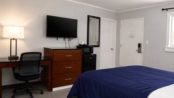 A hotel room with a bed, a desk with a chair, a dresser, a television, a mirror, and a door.