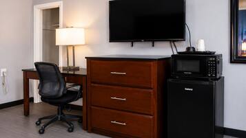 A hotel room with a flat-screen TV, a desk with a lamp, a black office chair, a wooden dresser, and a black refrigerator.