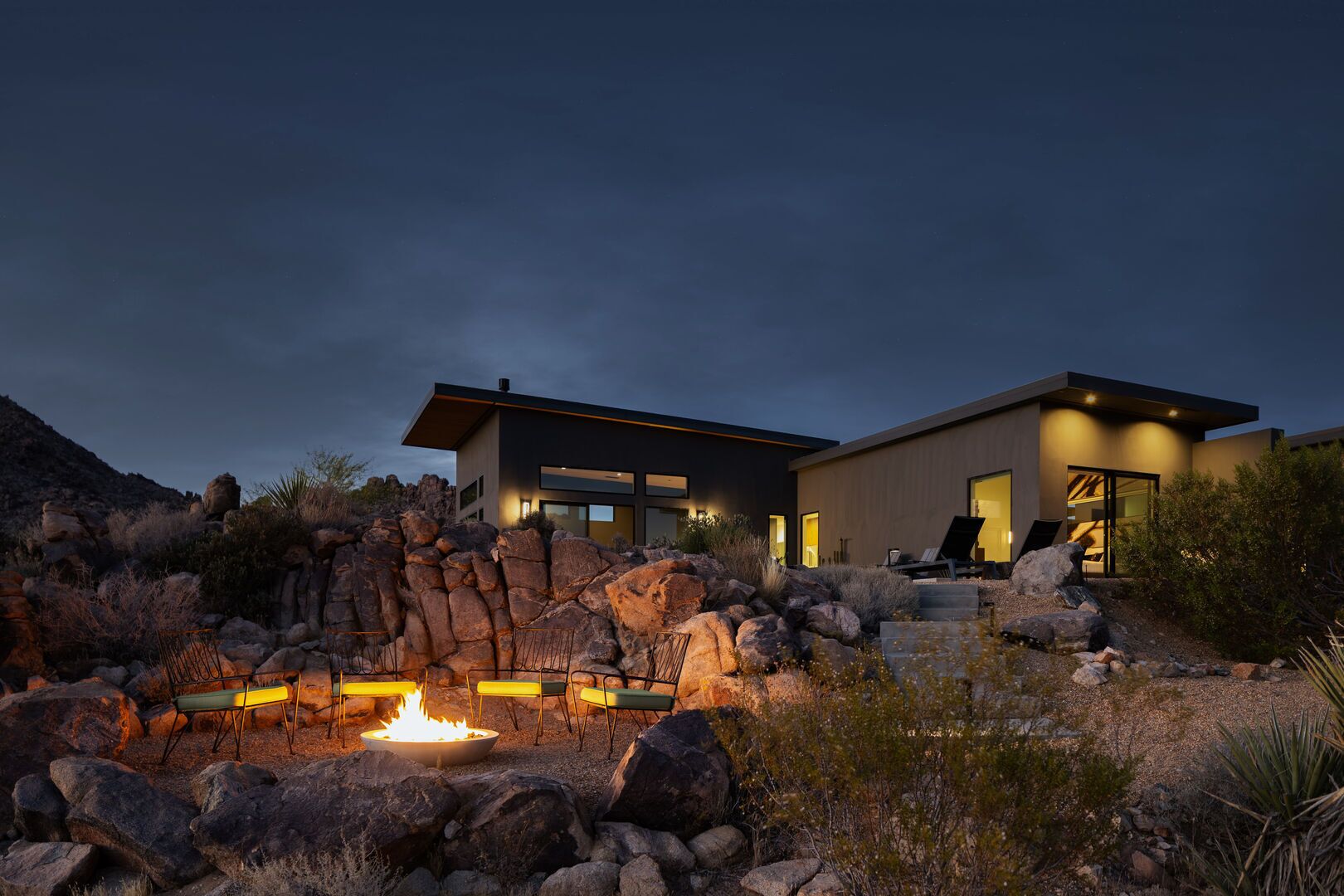 Une maison moderne à deux étages, avec un foyer au premier plan, entourée de rochers et de plantes typiques du désert.