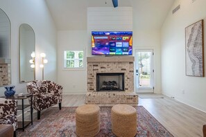 A living room with a fireplace, a sofa, ottomans, a TV mounted above the fireplace, and a large mirror.