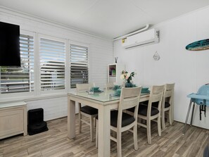 A dining area with a glass table, wooden chairs, and a blue vase with flowers. - Skye's Beach House | Downstairs, Shared Amenities | 2BR, Sleeps 6 | Spas (Malua Bay)