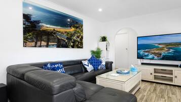 Ett modernt vardagsrum med en stor platt-TV, en svart lädersoffa och en tavla som föreställer ett kustlandskap.