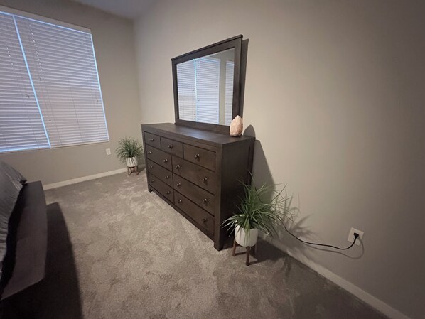 Un dormitorio con un aparador de madera oscura, un espejo, dos plantas en macetas, una ventana con persianas y piso alfombrado.