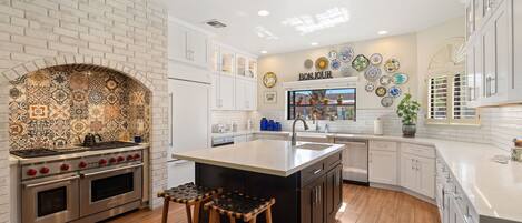 A kitchen with a white brick wall, a tiled backsplash, a large island with stools, and a 'BONJOUR' sign above the sink.
