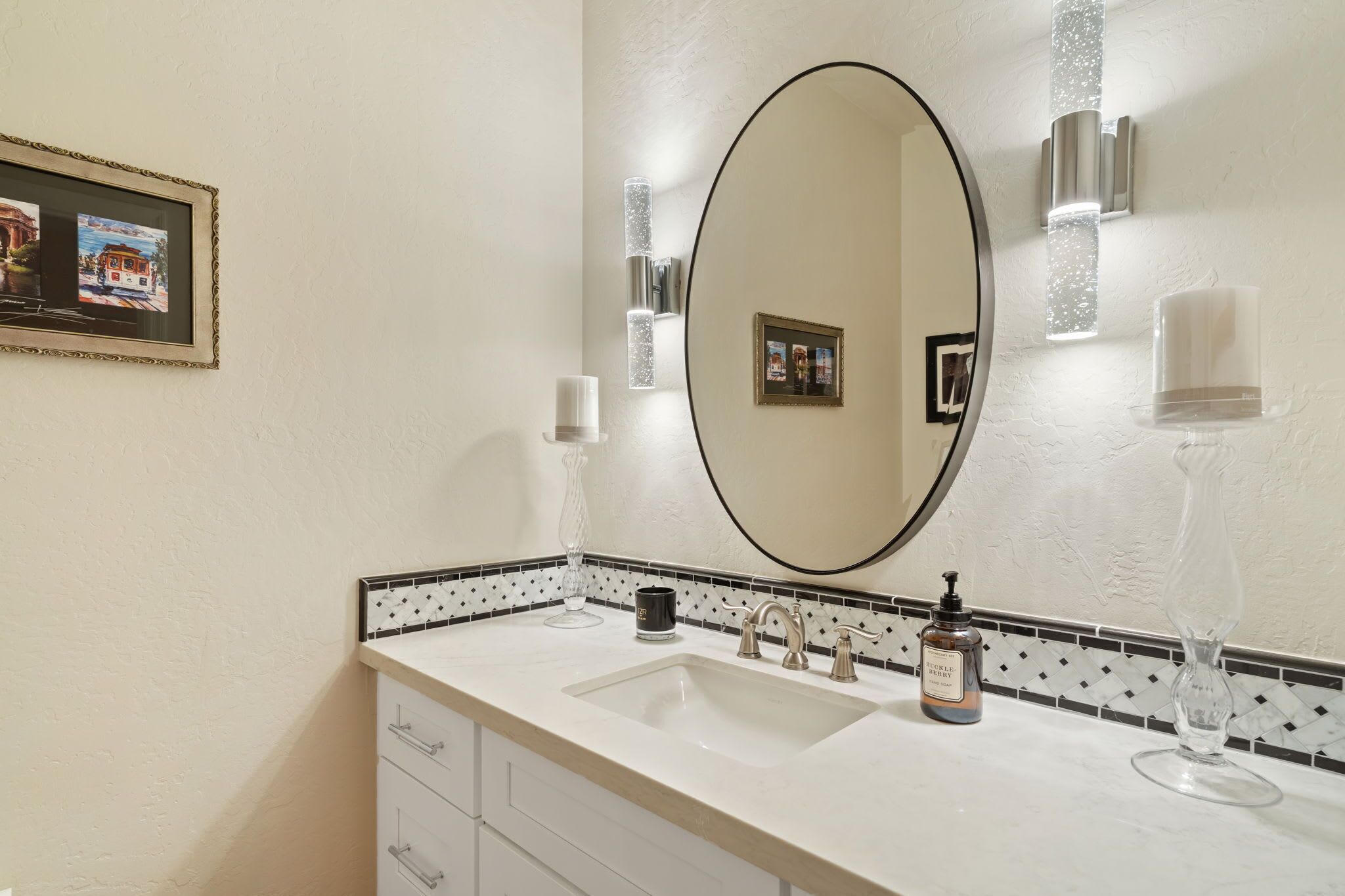 A bathroom with a large mirror, a sink, a bottle of hand soap, and a framed picture on the wall.