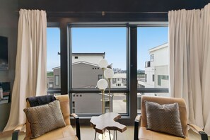 Une pièce moderne avec deux fauteuils en cuir, une petite table et un lampadaire. De grandes fenêtres offrent une vue sur les bâtiments voisins.