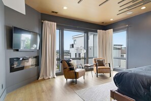 Une chambre d’hôtel moderne dotée d’un grand lit, de deux fauteuils, d’une télévision et offrant une vue sur la ville grâce à de grandes fenêtres.