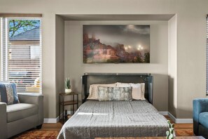 Un dormitorio con una cama, una mesita de noche, una planta en maceta, una ventana con persianas y un cuadro en la pared.