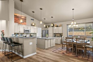 Una cocina moderna con una isla central, taburetes de bar, una zona de comedor con mesa y sillas de madera, iluminación colgante y un ventanal con persianas.