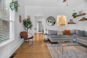 Uma sala de estar moderna com sofá, mesa de centro e uma poltrona. Há uma janela grande com persianas, uma planta e obras de arte nas paredes.