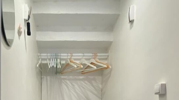 Um corredor estreito com parede branca, chão de madeira, espelho, cabideiro com roupas e interruptor de luz.
