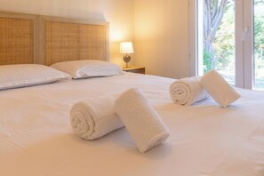 Ein Hotelzimmer mit einem großen Bett, zwei Kissen, einem Nachttisch mit einer Lampe und einem Fenster mit Blick auf Bäume.