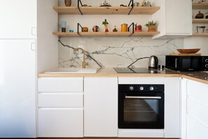 A kitchen with white cabinets, a marble countertop, a black oven, a microwave, and wooden shelves with various items.