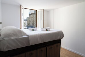 A neatly made bed with white linens and towels, a wooden headboard, and a view of a stone building through an open door.