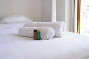 A hotel room with a neatly made bed, two rolled white towels, and a bottle of shower gel.