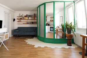 A living room with a sofa, a bookshelf, a dining area, and a kitchen visible through a green-framed glass partition.