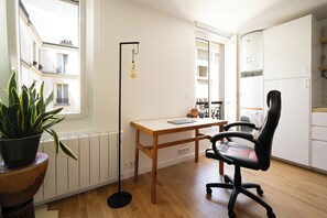 Une pièce avec un bureau en bois, un fauteuil de bureau noir, une plante en pot, un radiateur et une vue sur un bâtiment à l’extérieur.