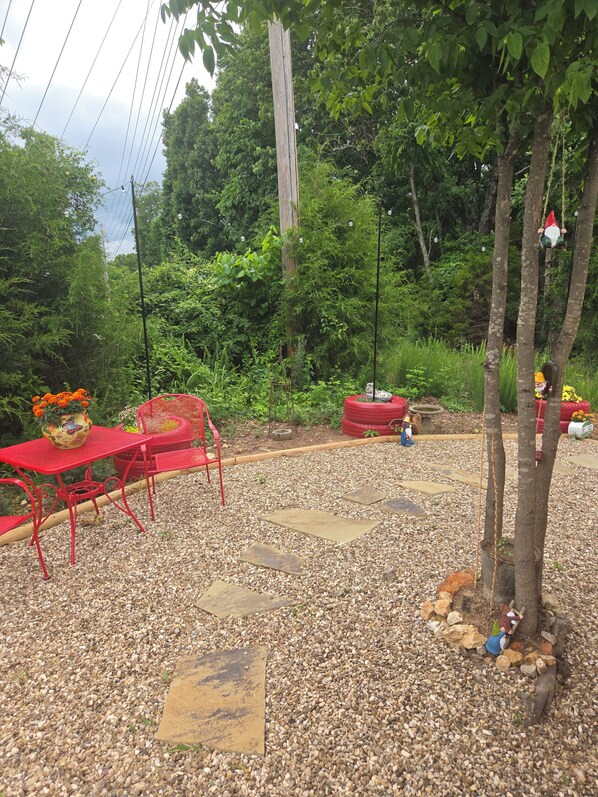 Un jardín con una mesa y sillas de metal rojo, un jarrón decorativo con flores y una estatua de gnomo.