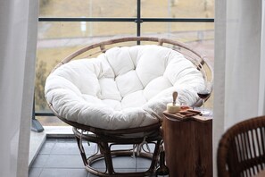 Ein Zimmer mit einem großen Fenster, ein Korbstuhl mit weißem Kissen sowie ein Holztischchen mit einer Flasche und Gläsern.