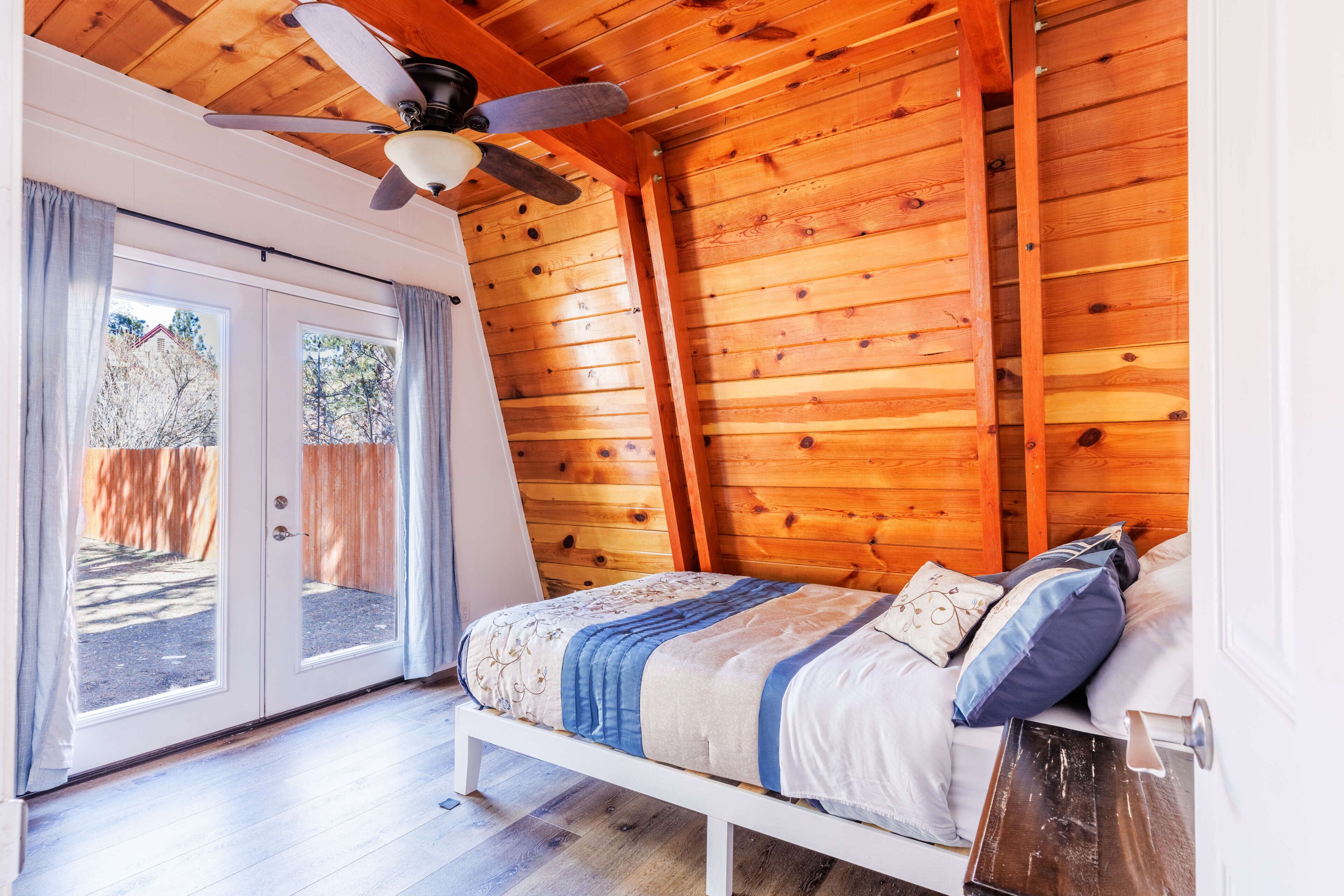 Un dormitorio con techo de madera, ventilador de techo, cama con cubrecama azul y blanco, y puerta corredera de vidrio que da al exterior.