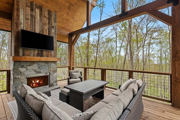 Eine Holzterrasse mit Kamin aus Stein, einem Flachbildfernseher und Sitzgelegenheiten im Freien.