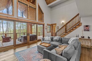Ein geräumiges Wohnzimmer mit einer großen, grauen Eckcouch, einer Holztreppe und großen Fenstern mit Blick auf den umliegenden Wald.