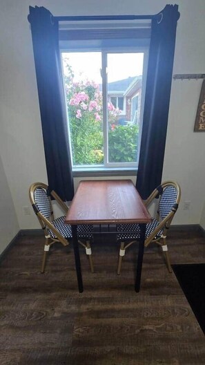 Quarto com mesa de madeira e duas cadeiras, janela com cortinas e vista para o verde lá fora.