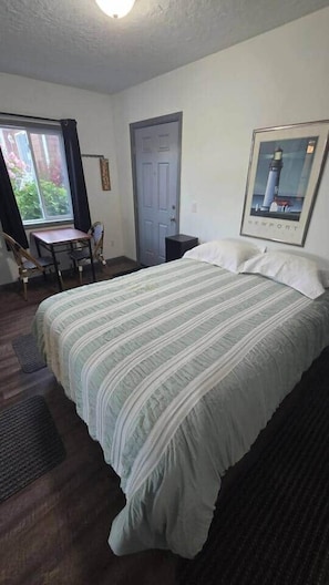 Quarto com cama, escrivaninha, cadeira, porta e quadro pendurado na parede.