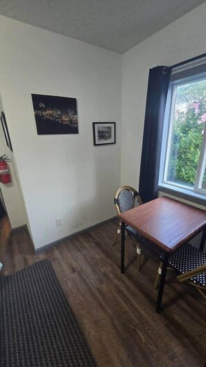 Um quarto com piso de madeira, uma mesa de jantar com cadeiras, uma janela com vista para o verde e duas quadros na parede.