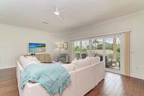 A spacious living room with a white sofa, a ceiling fan, a television, and a view of the outdoors through large sliding glass doors. - Still Waters hosted by Beach Retreats FL (Holmes Beach)