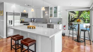 A modern kitchen with white cabinetry, stainless steel appliances, a large island with stools, pendant lighting, and a view of the outdoors. - Sol y Soledad: A Modern Palm Springs Retreat with Private Pool, Spa & Office (Palm Springs)