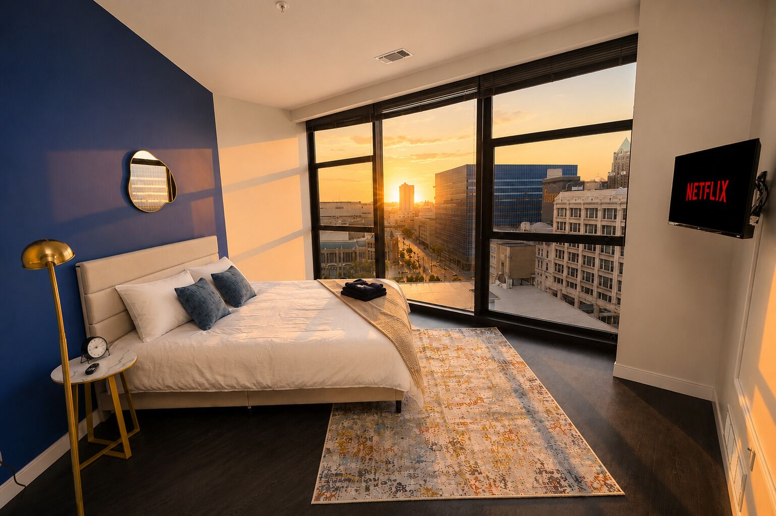 A modern hotel room with a large bed, a view of the cityscape, a flat-screen TV, and a unique wall mirror.