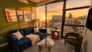 A modern living room with a blue sofa, a glass coffee table, and a view of a cityscape at sunset. - Stylish Westown 2BR Retreat 10 min to Fiserv Forum (Milwaukee)