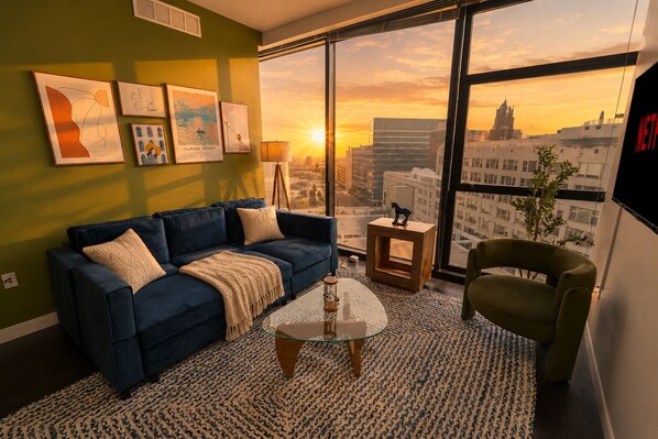 Een moderne woonkamer met een blauwe bank, een glazen salontafel en uitzicht op een stadsbeeld bij zonsondergang.